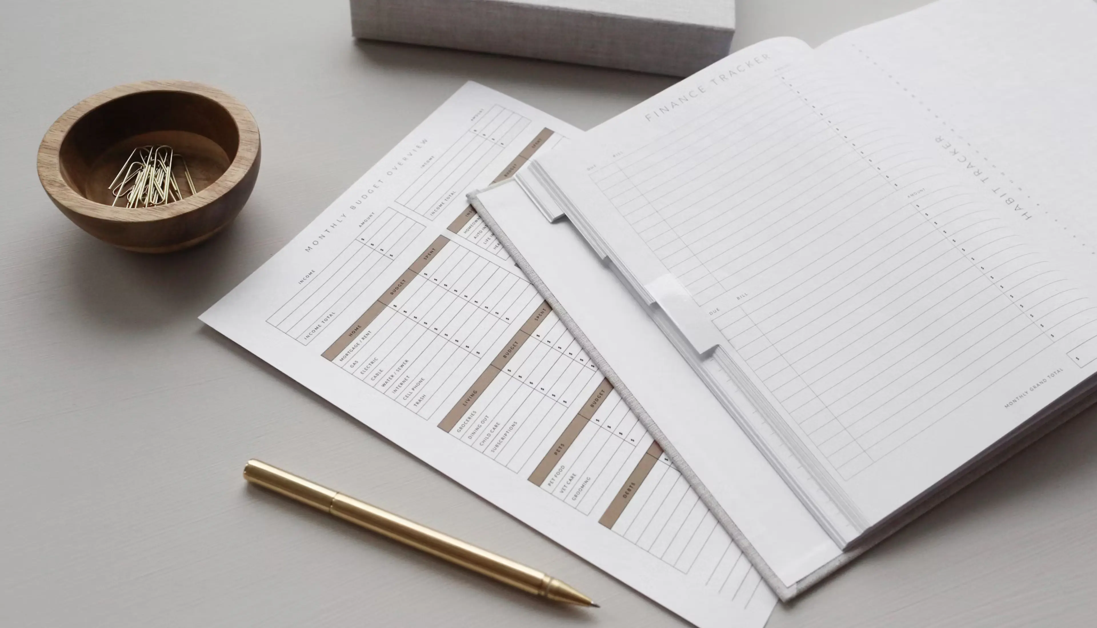 Monthly budget planning sheets and a notebook on a table with a gold-coloured pen and paperclips in a wooden holder.
