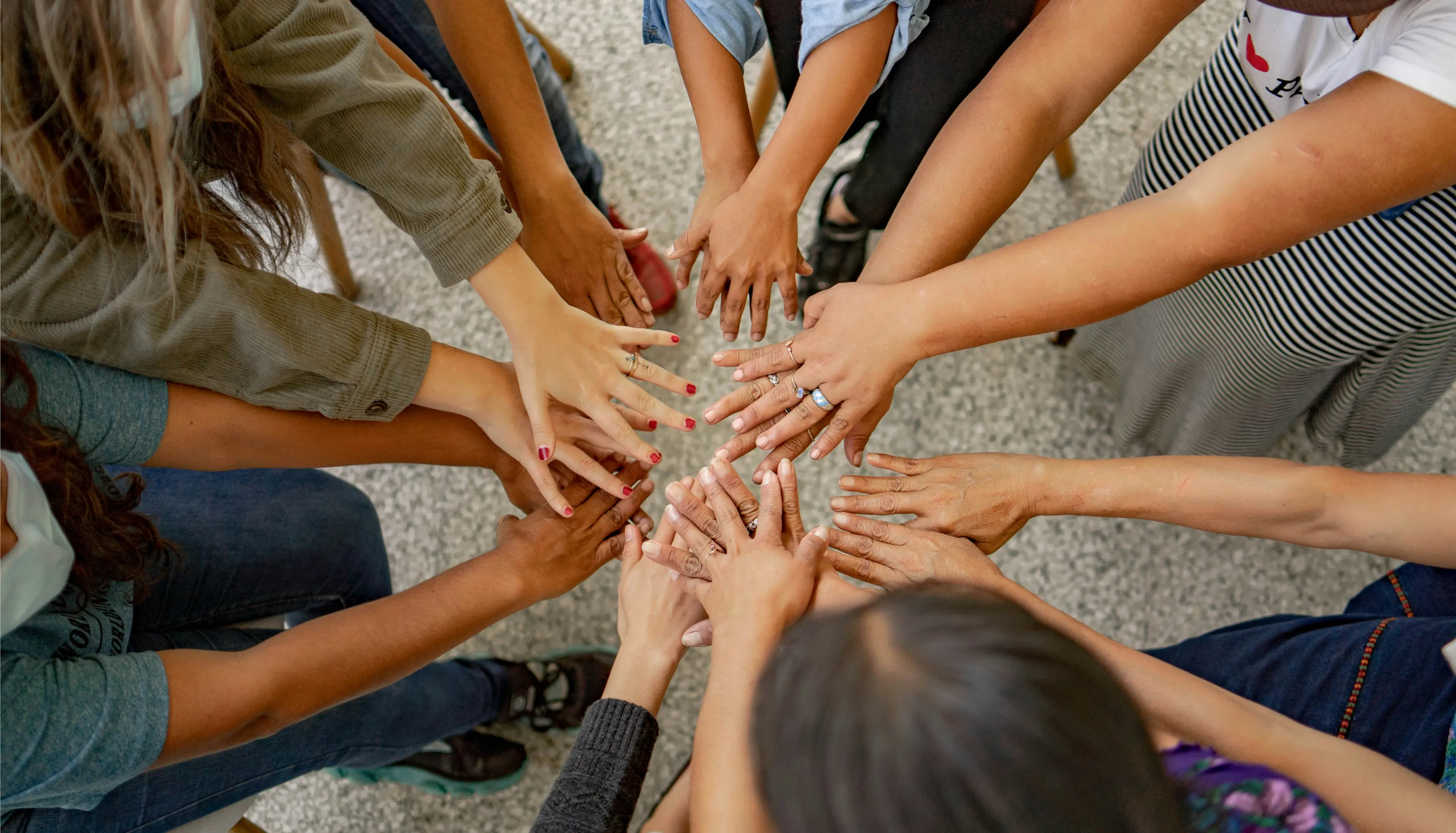 A diverse group of people stand in a circle, extending their hands toward the centre with various skin tones, nail colours, and jewellery visible, symbolising teamwork and unity.