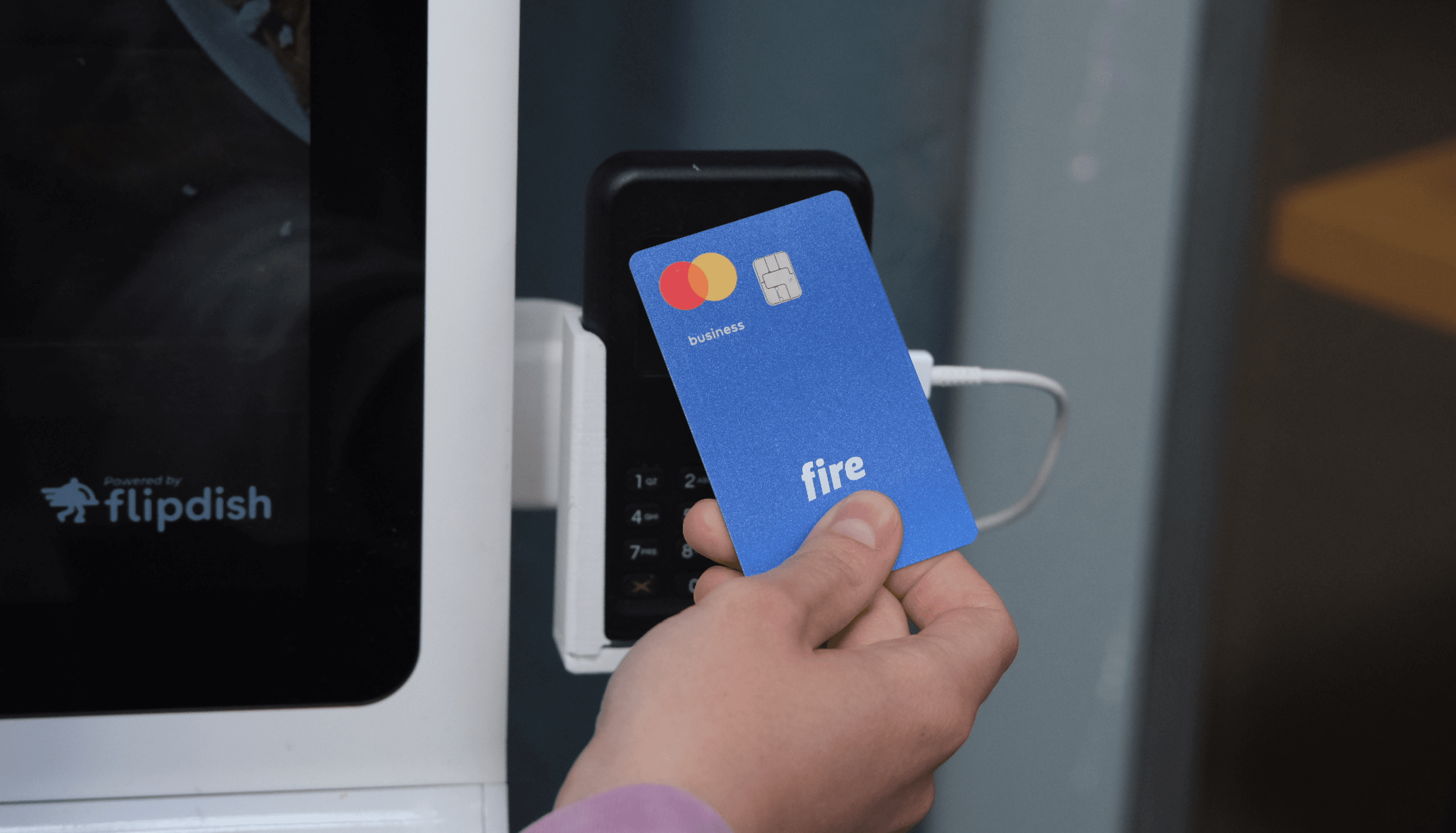 A close-up of a person’s hand making a payment by tapping a blue Fire business debit card on a POS terminal.
