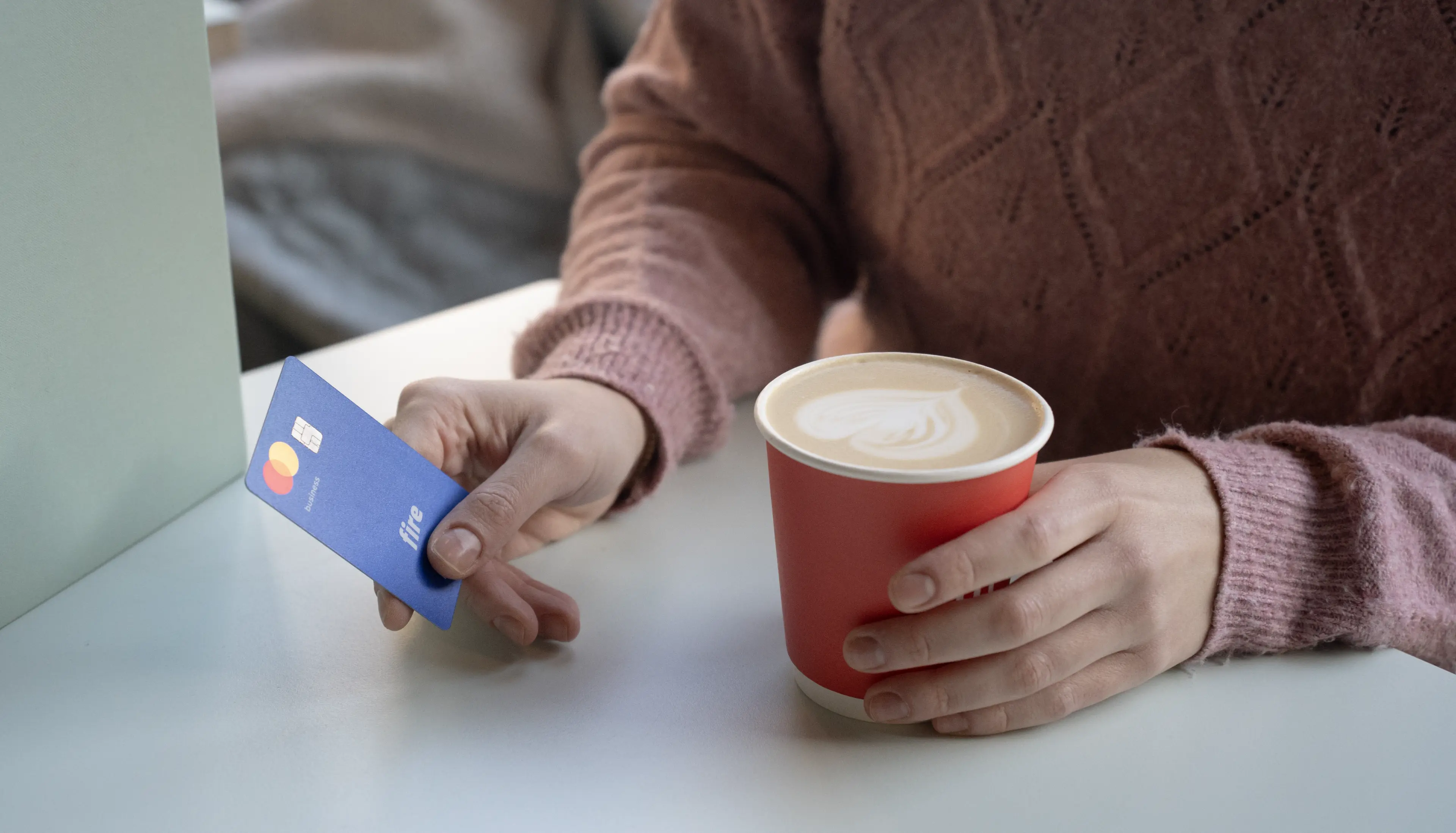 Close-up of a person holding a Fire business debit card in one hand and a paper coffee cup in the other, dressed in a pink jumper. The image suggests the act of tipping while purchasing a coffee.