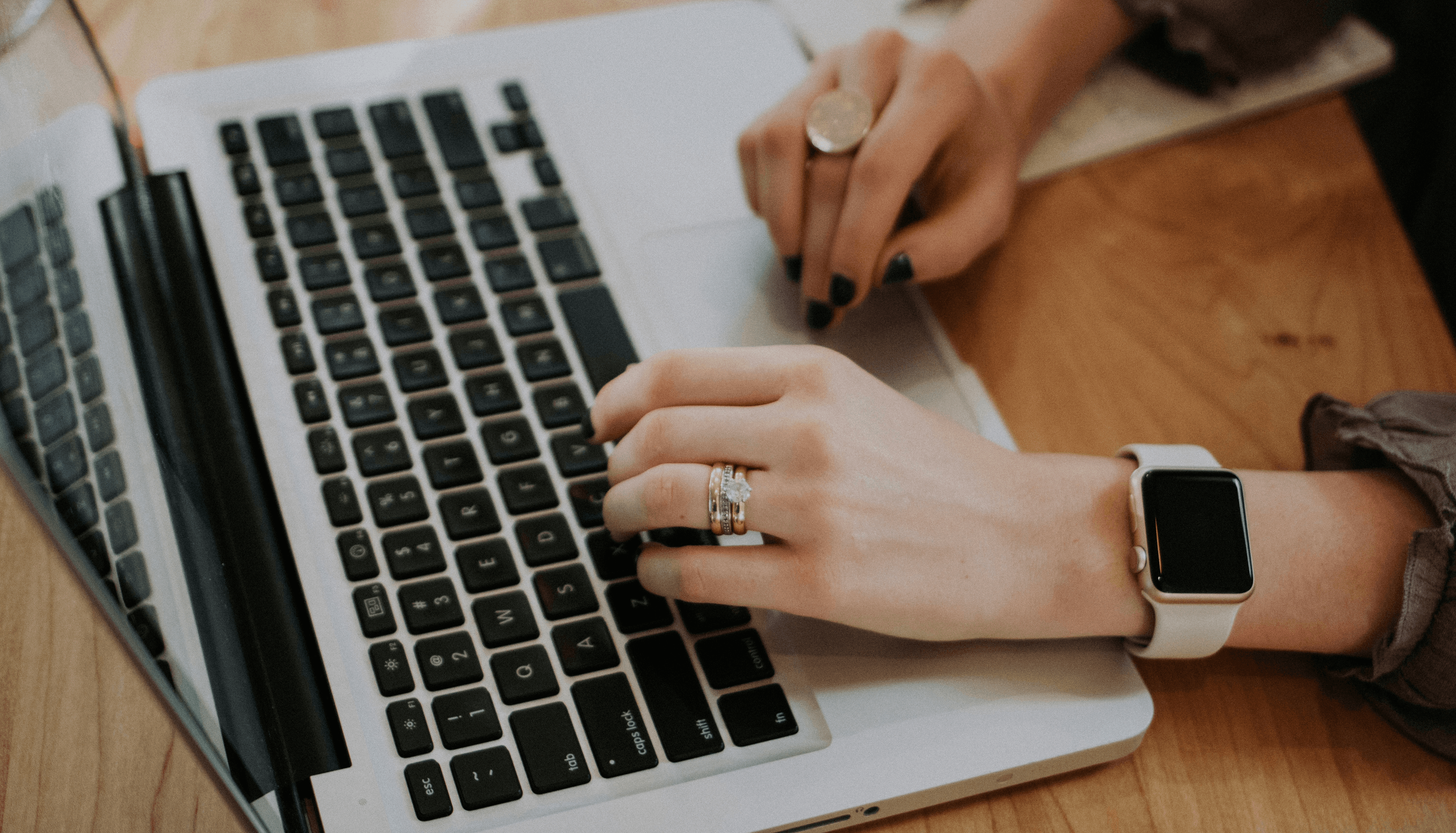 Close-up of a woman’s hands with black-painted nails, wearing multiple rings and an Apple Watch, typing on a computer.