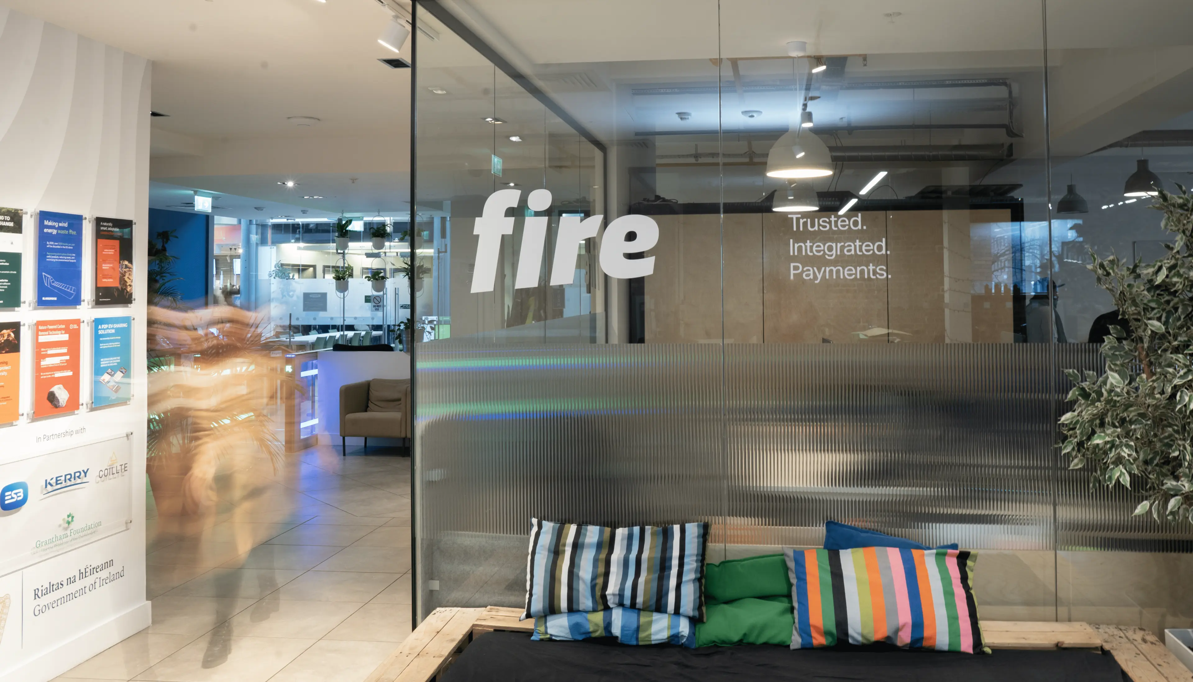 Corner of Fire’s glass office in Dogpatch Labs, featuring colourful pillows in the foreground, with the Fire logo and the tagline "Trusted. Integrated. Payments." displayed on the large glass walls.
