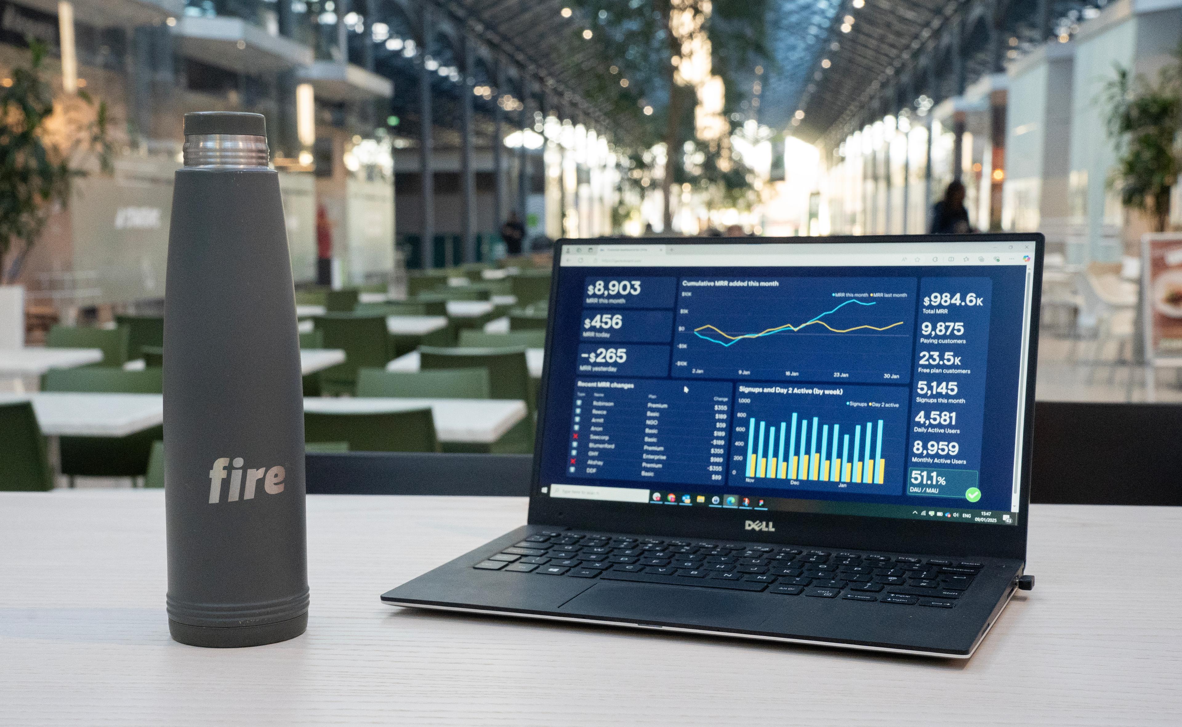 A Fire-branded water bottle and a laptop displaying financial charts on a table in a spacious, well-lit hall with desks in the background.