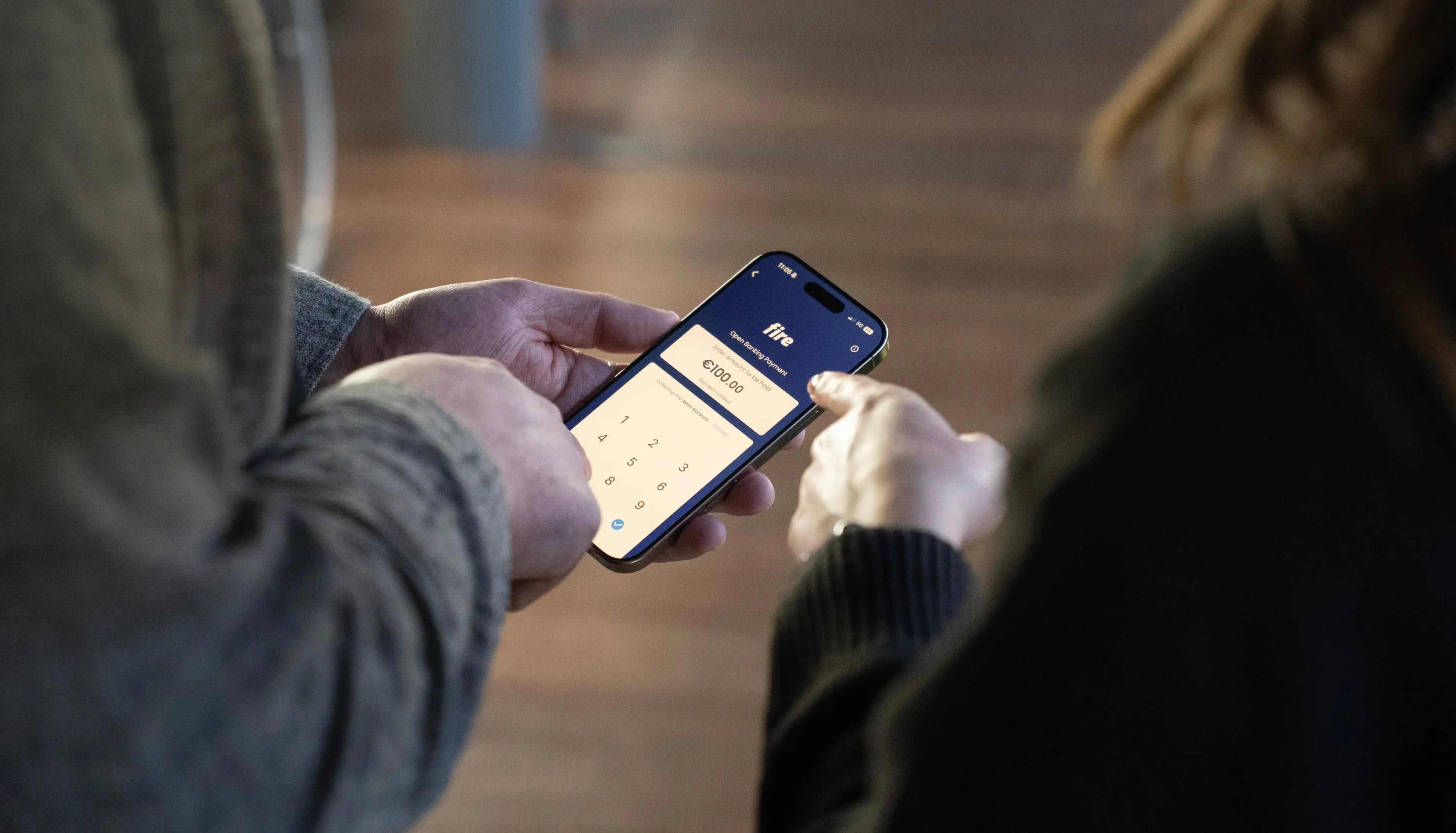 Close-up of two people's hands, one holding a mobile phone and the other pointing at a screen displaying a Fire open banking payment. The interaction illustrates a multi-user authorisation process, where individuals have different user roles and access levels, reviewing and approving transactions collaboratively.