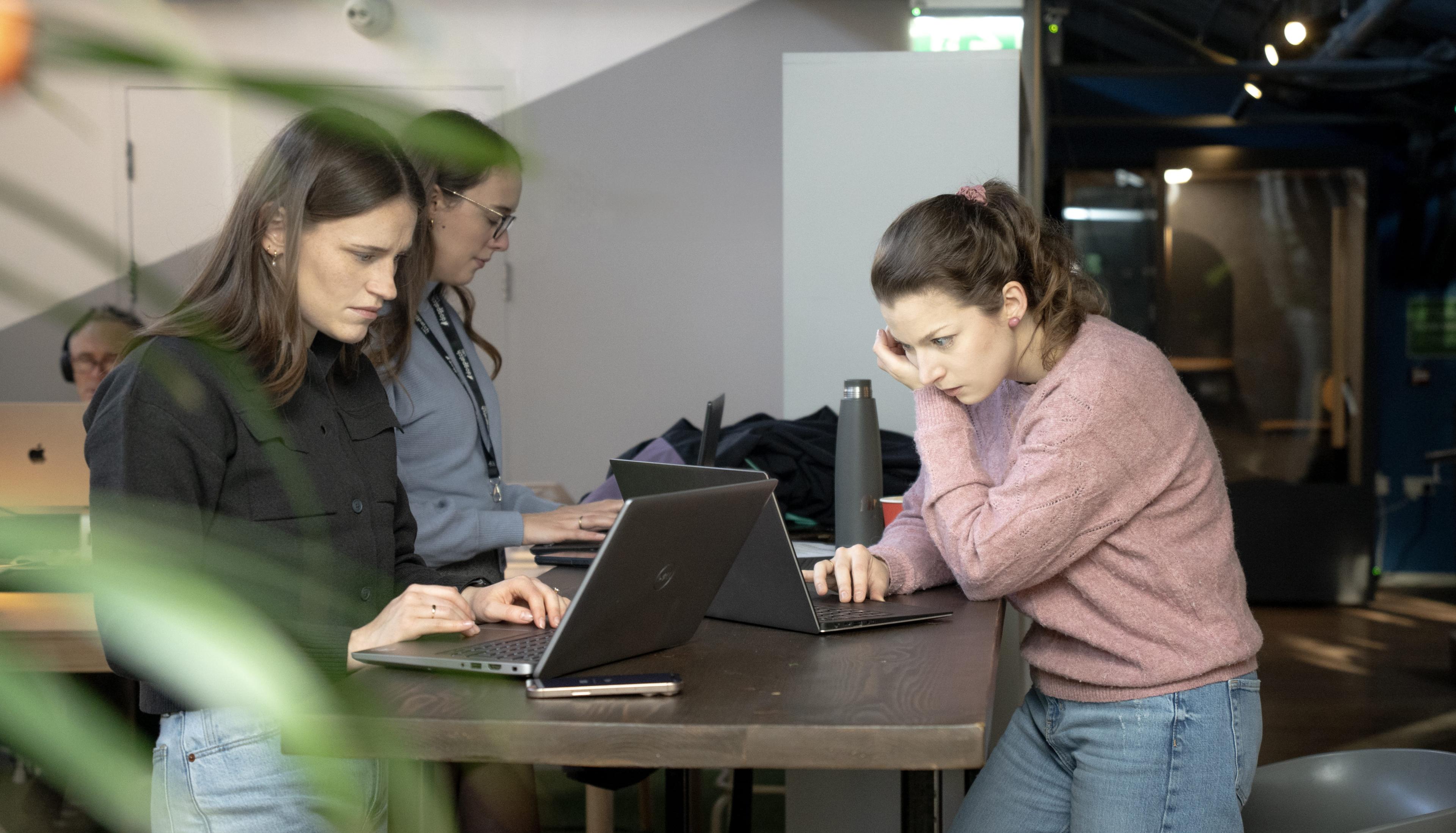 Three women working on laptops at a shared high table in a modern office space. Two are standing while one leans forward intently, suggesting focused collaboration. A plant is partially visible in the foreground.