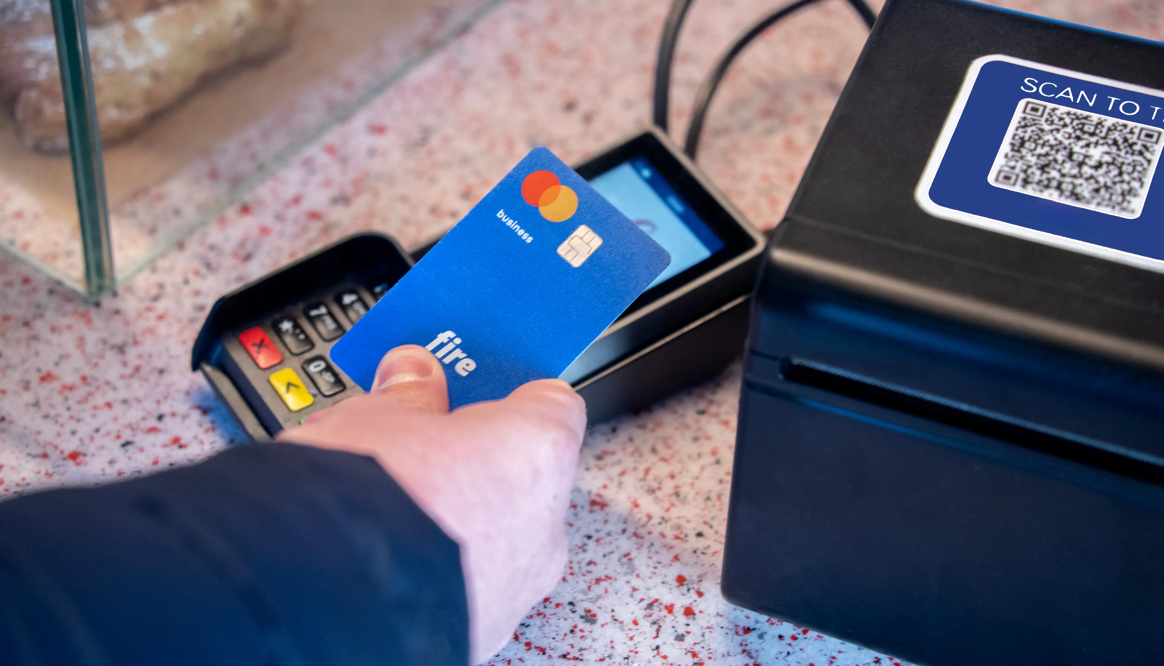 Close-up of a person’s hand, paying with a blue Fire business debit card at a POS terminal in a coffee shop, illustrating business debit card spend tracking.