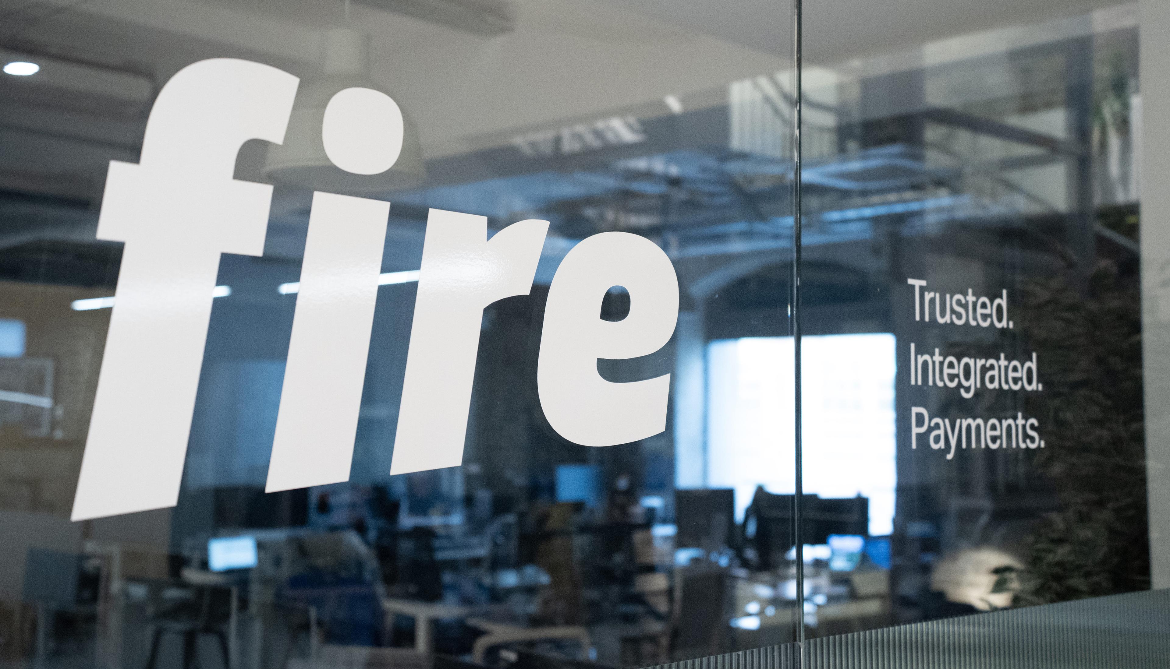 Close-up of the Fire logo and the tagline “Trusted. Integrated. Payments.” displayed on the glass wall of Fire’s office, with a view of a modern workspace and desks in the background.