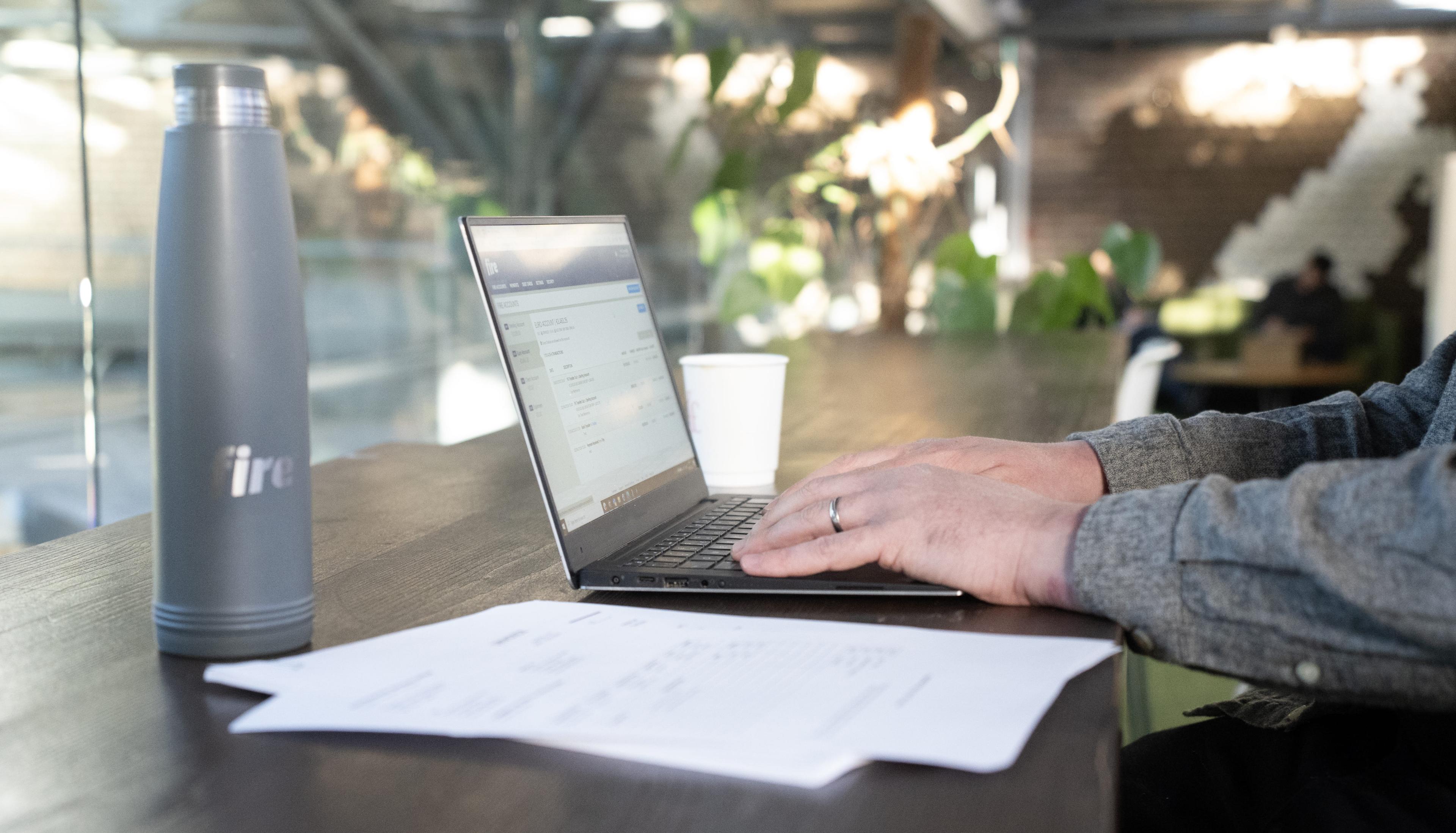 A person is working on a laptop displaying the Fire web app, seated at a high table in a modern, sunlit workspace. Printed documents, a grey Fire-branded water bottle, and a takeaway coffee cup are on the table. The background is well-lit with natural light and features greenery.