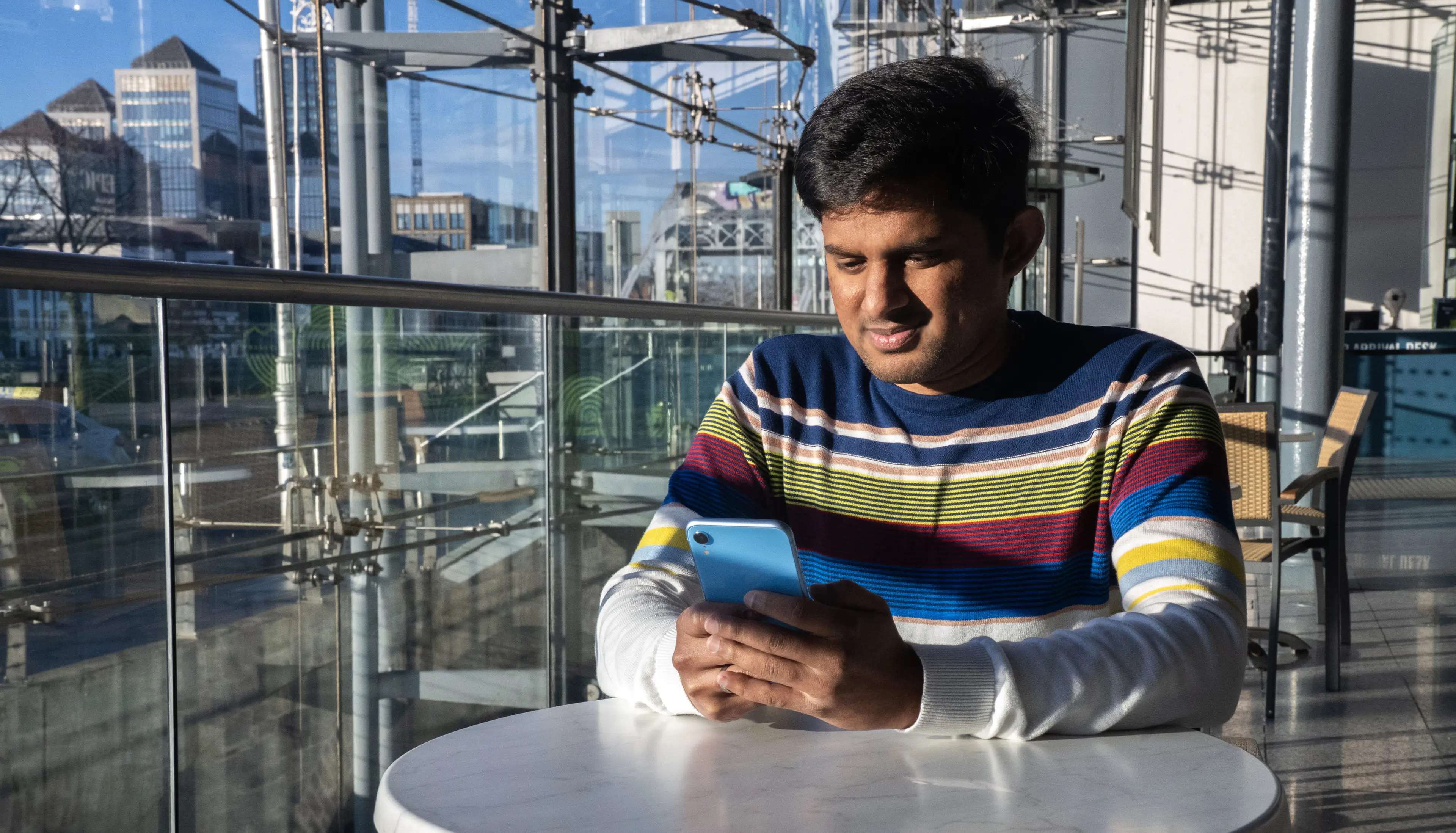 A man sits at a round white table in a modern glass-walled building, looking at a smartphone in his hands. He is wearing a dark blue sweater with colourful horizontal stripes. The background shows an urban landscape with high-rise buildings and metal structures.