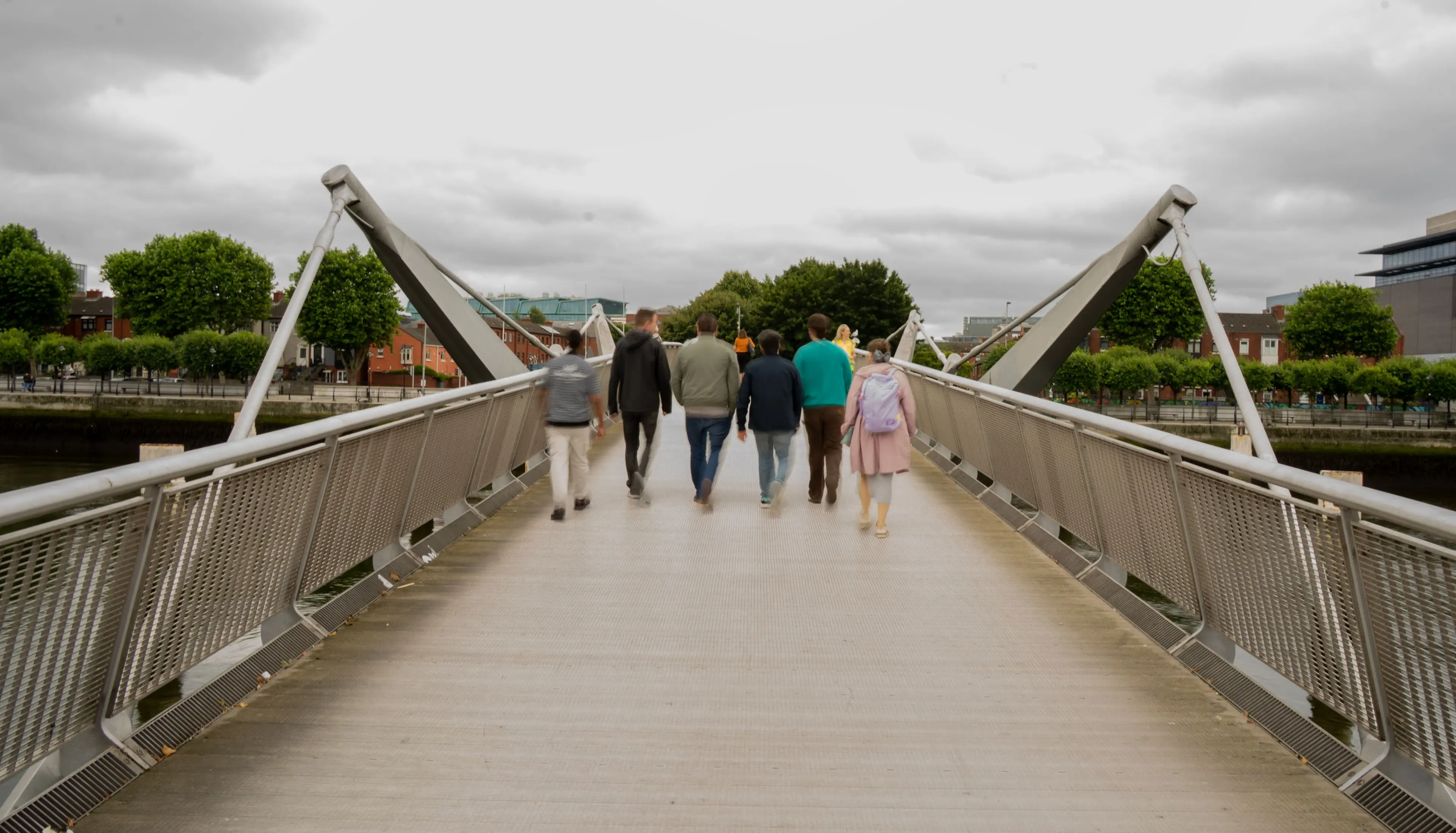 Group of people walking across a modern pedestrian bridge over a river, with trees and buildings in the background under a cloudy sky.