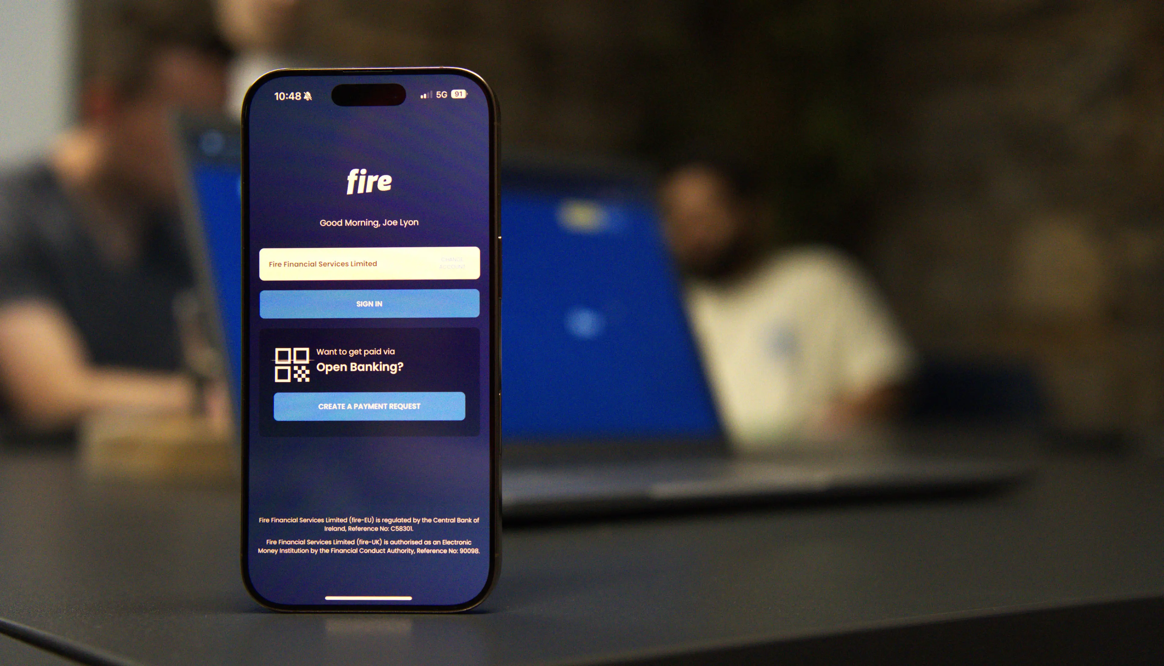 A smartphone on a desk displays the Fire app login screen, showing the option to create an open banking payment request. The screen reads “Want to get paid via Open Banking?” with a button below saying “Create a payment request.” In the blurred background, two people sit at a table with laptops.