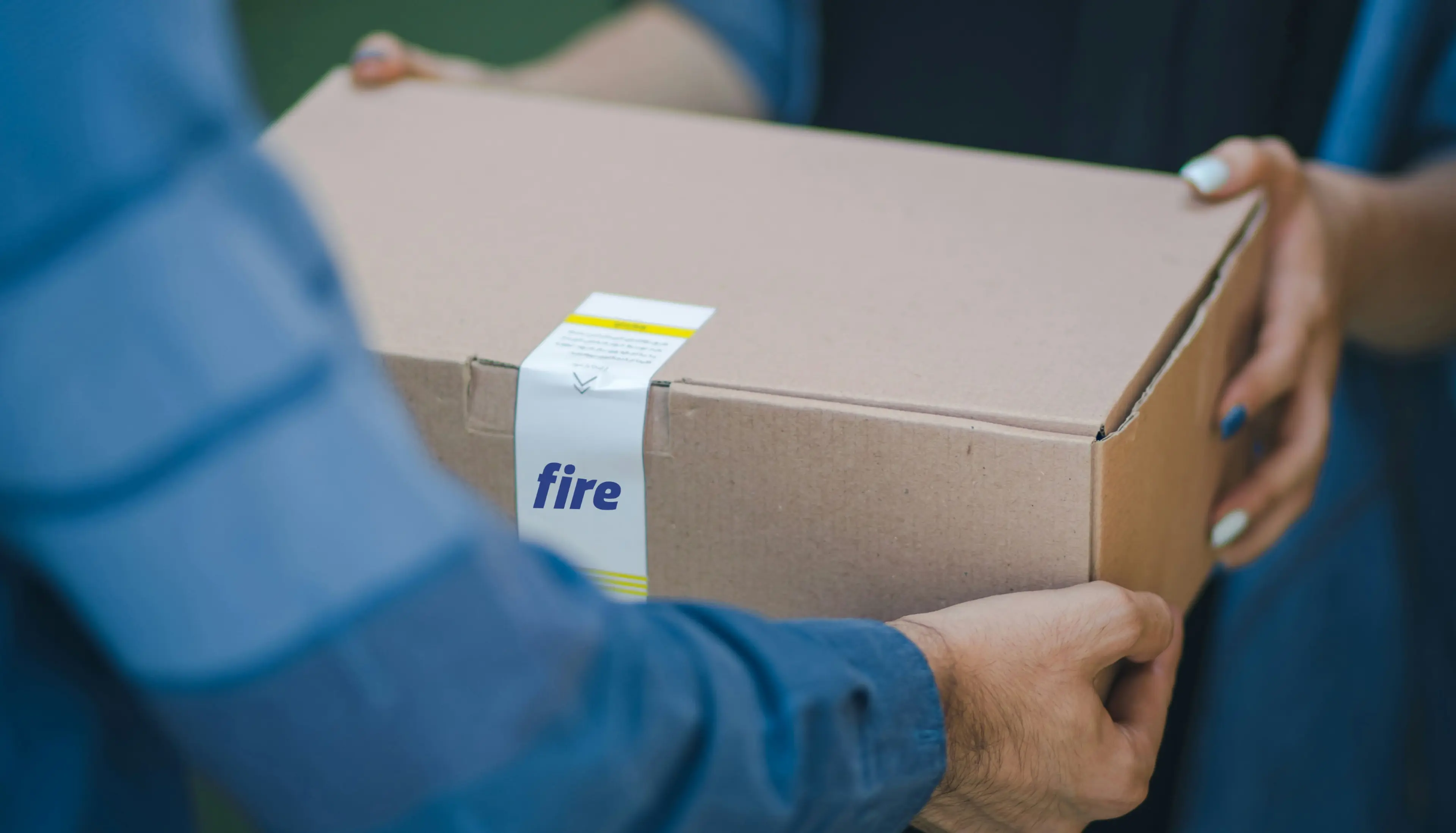 Close-up of two people’s hands: a courier in a blue jacket handing a cardboard delivery box with the Fire logo to a person with painted nails.