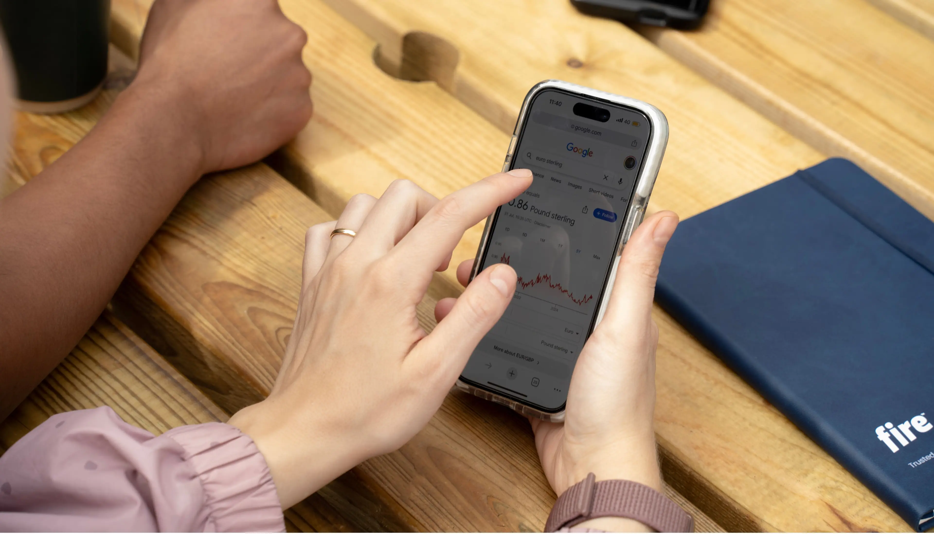 Close-up of hands at a wooden desk, one holding a smartphone displaying a euro–sterling currency fluctuation chart and the other pointing at the screen, with a Fire-branded notepad visible on the desk.