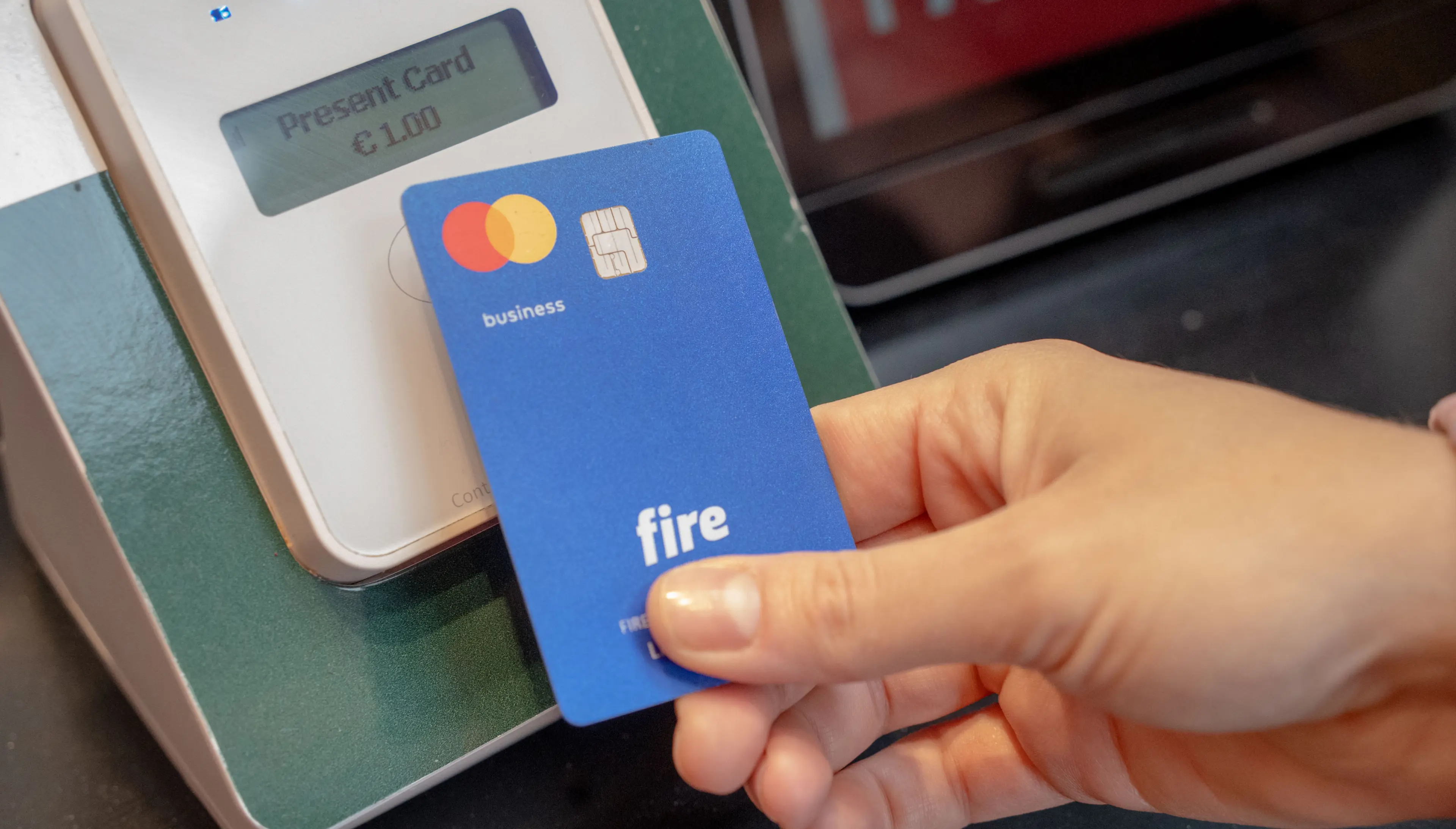 A person holds a blue Fire business debit card close to a card reader to make a contactless payment.