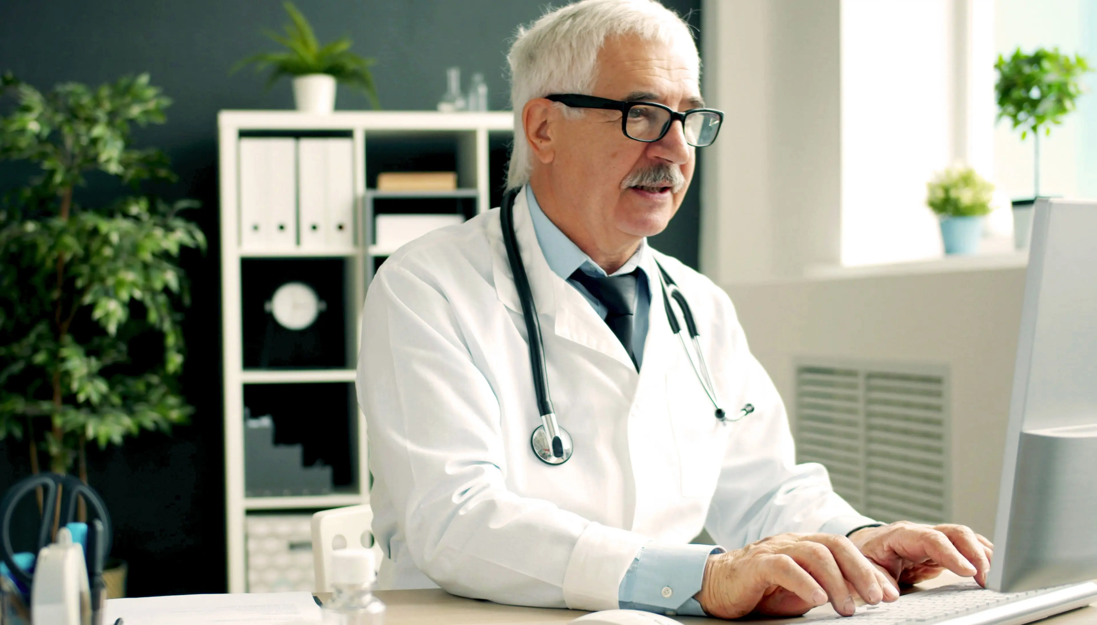 A doctor sitting at a desk typing on a computer in a medical office.
