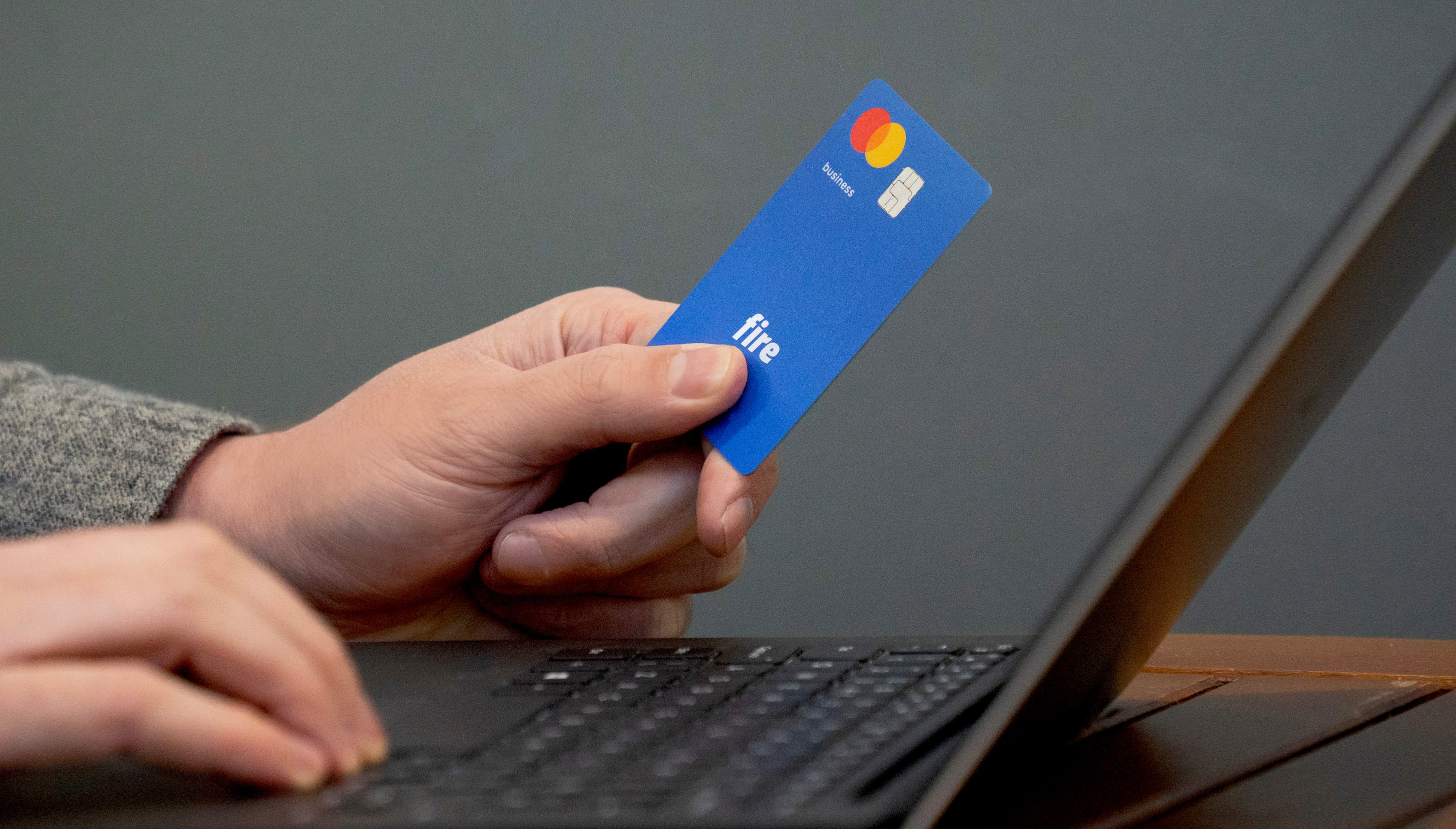 A hand holding a blue Fire Mastercard business debit card beside a laptop.
